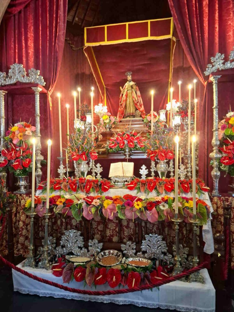 San Marcos Evangelista está de pie en su altar, rodeado de flores y velas encendidas. Un libro abierto se encuentra a sus pies. El altar está adornado con manteles brocados en tonos rojos y dorados. Los candelabros de plata están ubicados a ambos lados y están llenos de velas. El fondo es de color rojo y hay cortinas de color rojo. Todo el altar está engalanado para la Romería de Tegueste.