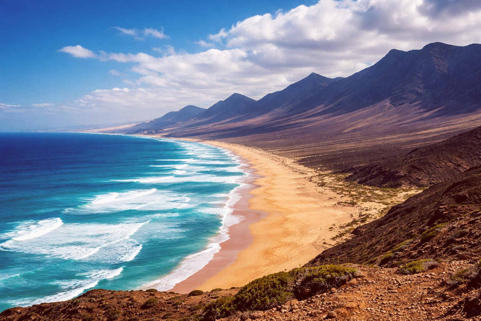 Vista panorámica horizontal de la Playa de Cofete en Fuerteventura con el Atlántico azul intenso, olas rompiendo en la orilla dorada y las montañas de Jandía al fondo bajo cielo parcialmente nublado.